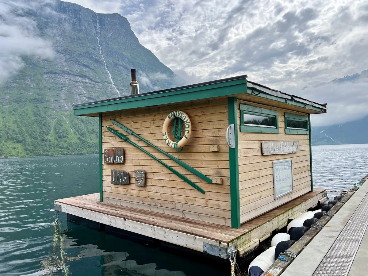 A wooden sauna perched above a Norwegian fjord with mountains reflected in still water